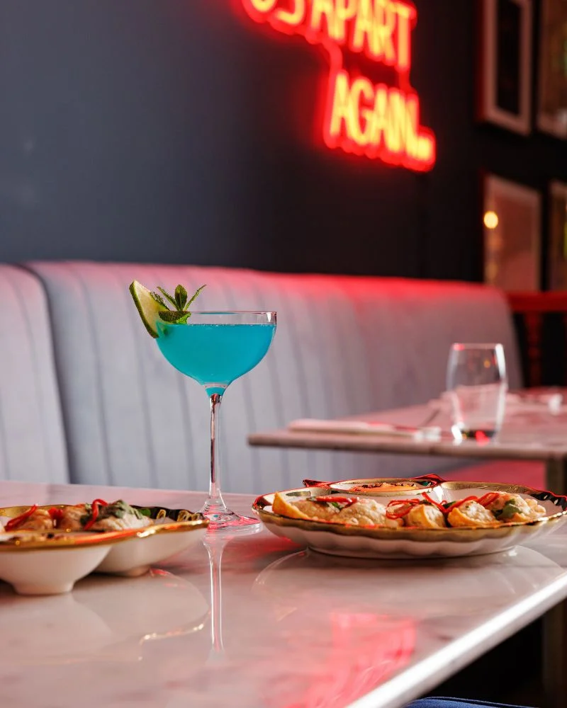 A striking blue cocktail on a marble table with a backdrop of neon 'Do It Again' sign, complementing an appetizing Asian cuisine dish in a Dublin eatery.